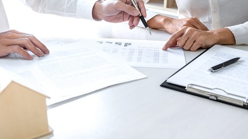 Zwei Menschen sitzen am Schreibtisch und schauen sich Dokumente an. Auf dem Tisch steht ein kleines Modellhaus aus Holz