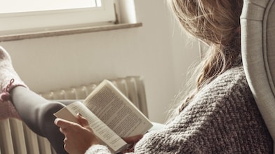 Frau sitzt in einem Sessel und liest ein Buch. Die Füße legt sie auf einer Heizung ab.