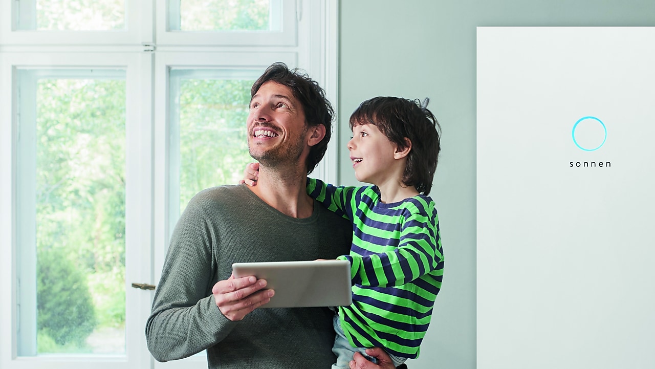 Ein Mann steht in der Küche, trägt sein Kind und hält ein Tablet in der Hand. Im Hintergrund befindet sich eine sonnen Batterie