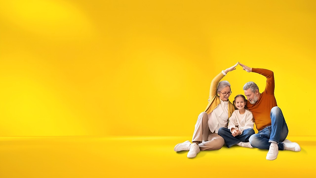 Familie sitzt zusammen fröhlich auf dem Boden und formt mit den Händen symbolisch ein Haus, vor einem leuchtend gelben Hintergrund. 