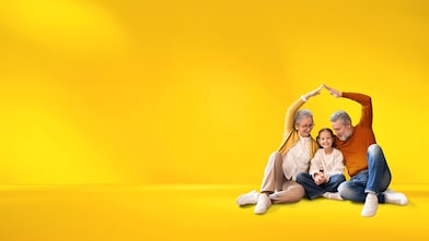 Familie sitzt zusammen fröhlich auf dem Boden und formt mit den Händen symbolisch ein Haus, vor einem leuchtend gelben Hintergrund. 