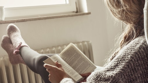 Eine Frau liest ein Buch in einem Sessel und ihre Füße sind auf der Heizung abgelegt.