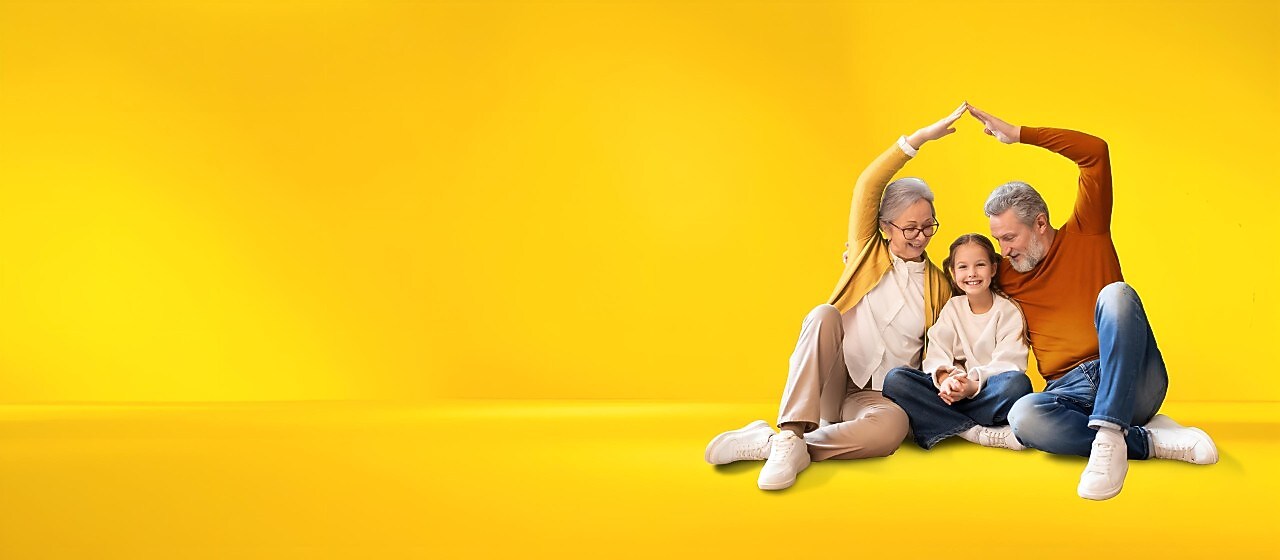 Familie sitzt zusammen fröhlich auf dem Boden und formt mit den Händen symbolisch ein Haus, vor einem leuchtend gelben Hintergrund. 