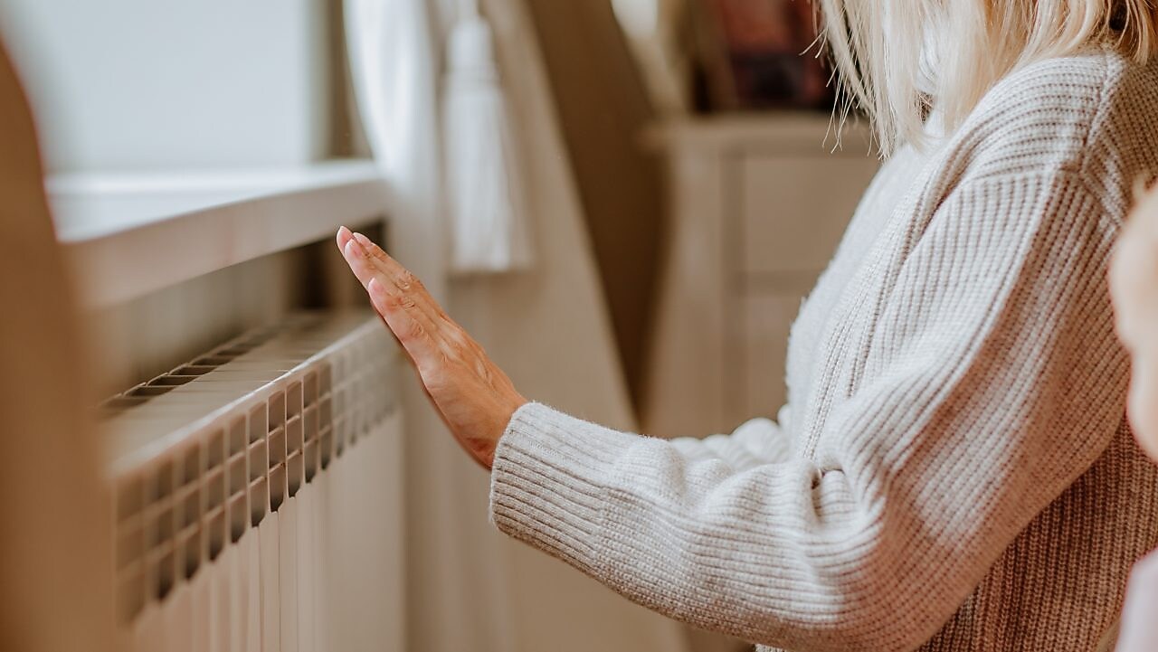 Eine Frau in einem beigen Winterpullover steht in der Nähe der Heizung und wärmt sich die Hände 