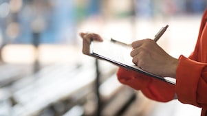Person in orangener Kleidung schreibt mit einem Stift auf ein Klemmbrett