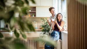 Paar steht mit Getränken am Fenster einer hellen, modernen Küche mit vielen Zimmerpflanzen.