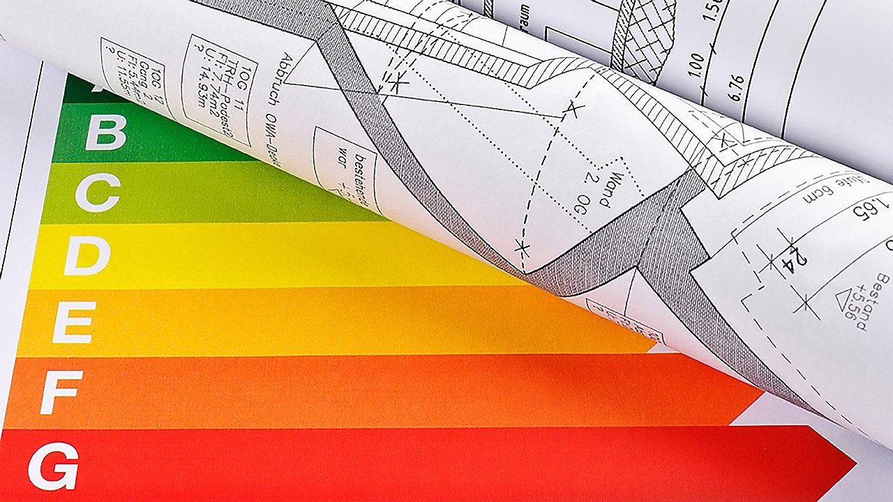 Gerollter Bauplan liegt auf einem farbigen Energieeffizienzlabel mit den Klassen A bis G.