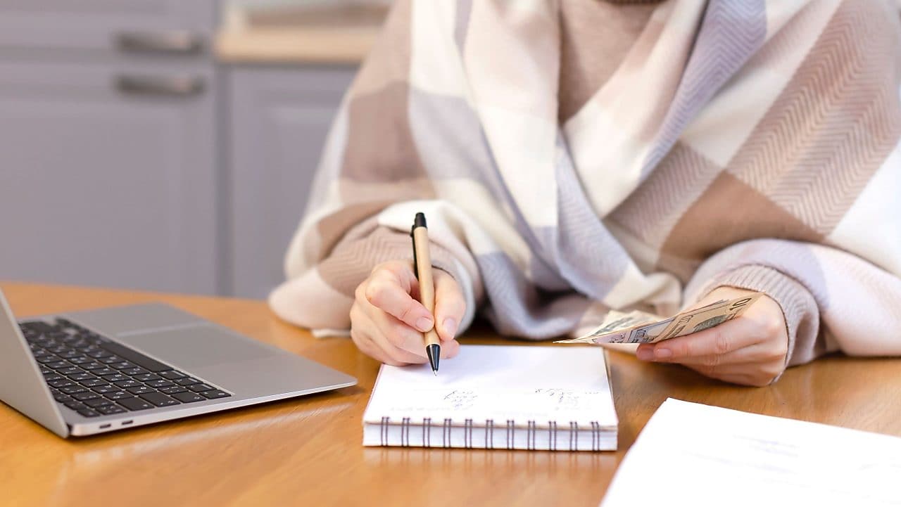 Eine Frau, eingehüllt in einen warmen Schal, zählt Geld und schreibt in ein Notizbuch.