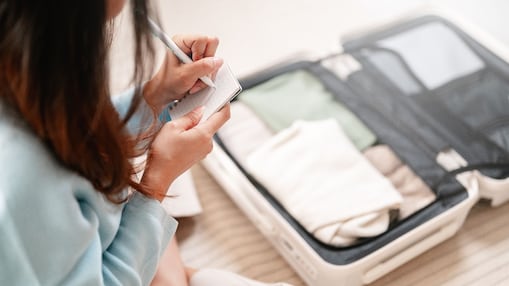 Frau packt einen offenen Koffer mit Kleidung und schreibt mit einem Stift Notizen auf ein Blatt Papier.