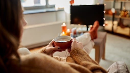 Eine Frau sitzt gemütlich zu Hause, hat die Füße auf dem Tisch, schaut fern und trinkt eine Tasse heiße Schokolade