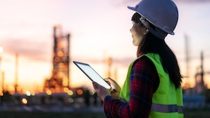 Eine Frau steht bei Sonnenuntergang mit Schutzkleidung und einem Tablet in der Hand im Außenbereich einer Raffinerie.
