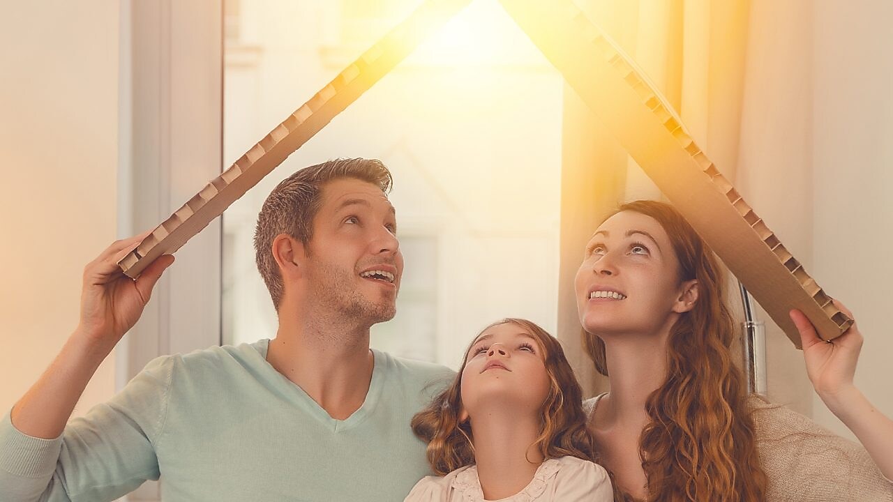 Ein Mann, Frau und ein Kind sitzen in einem Haus, halten sich ein Karton über den Kopf, das aussieht wie ein Dach. Die Sonne scheint hindurch.