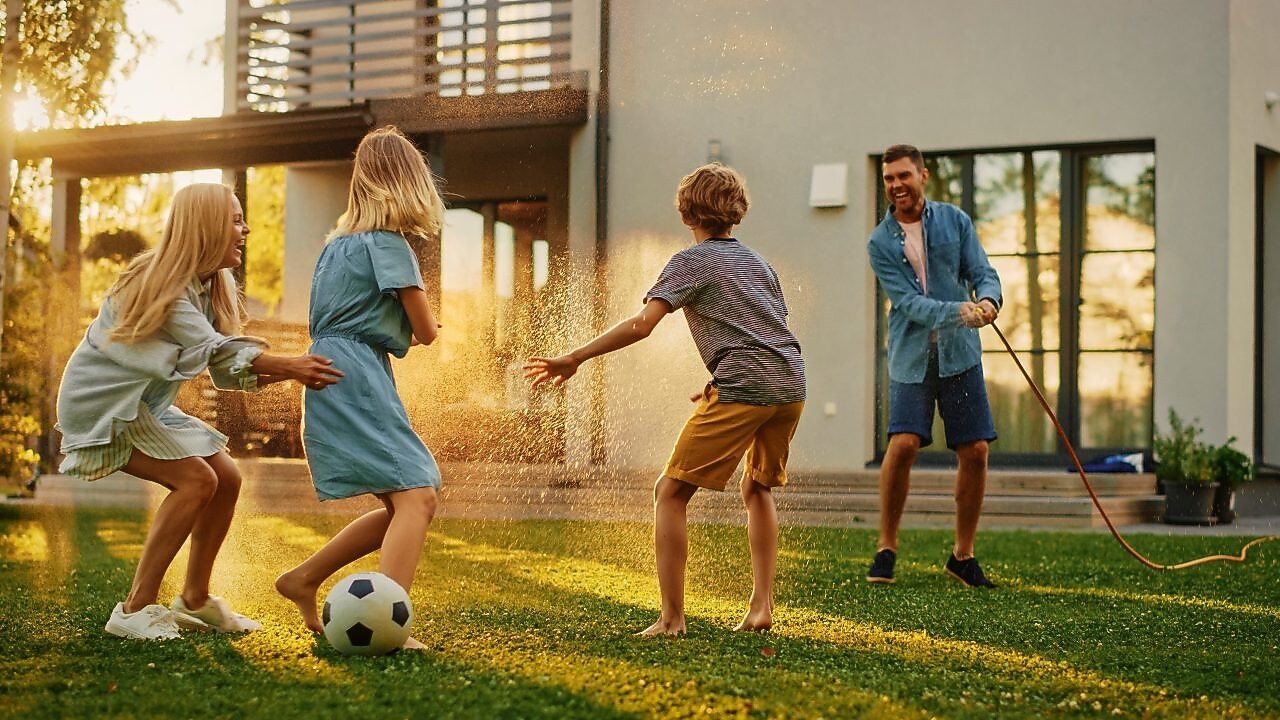 Eine Familie spielt draußen in ihrem Garten mit einem Ball und der Vater besprüht die Kinder mit Wasser aus dem Gartenschlauch