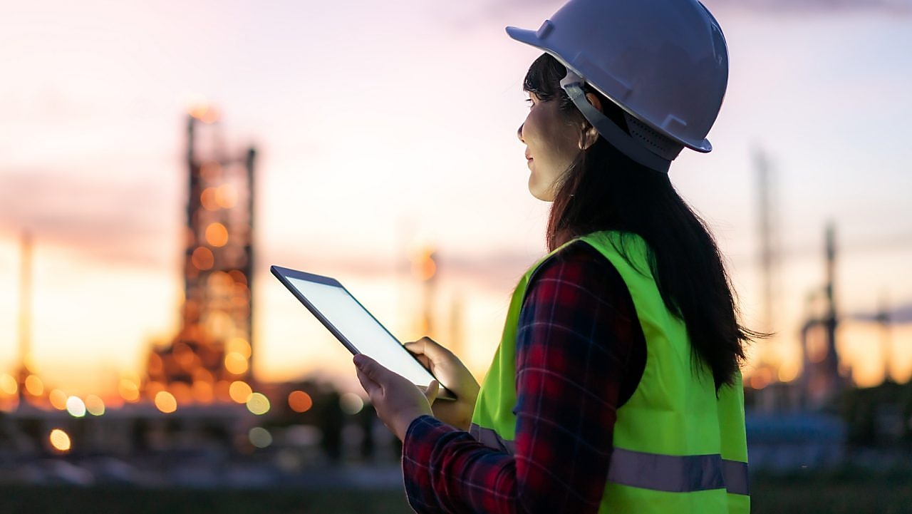Eine Frau steht bei Sonnenuntergang mit Schutzkleidung und einem Tablet in der Hand im Außenbereich einer Raffinerie.