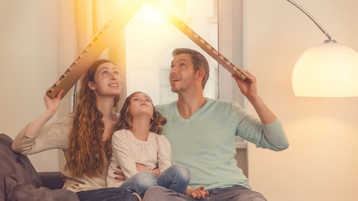 Familie sitzt auf dem Sofa und hält ein Dach aus Karton über ihre Köpfe, warmes Sonnenlicht fällt von oben ins Wohnzimmer
