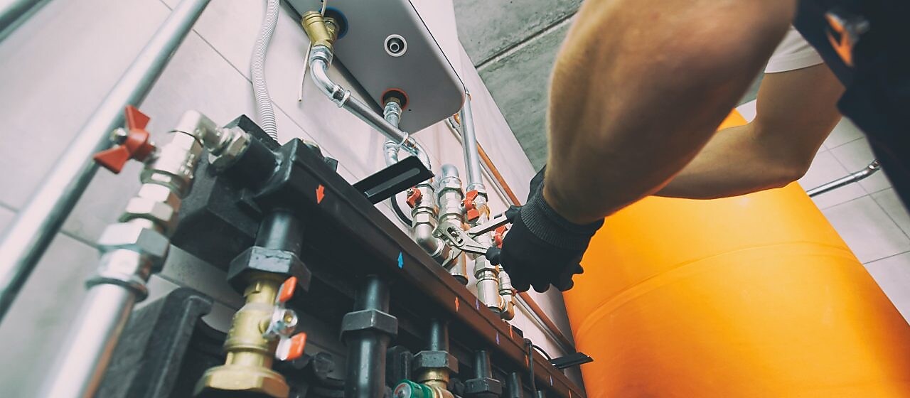 Jemand in einem Blaumann ist im Heizungsraum und verstellt etwas mit einer Rohrzange, in der Nähe eines Heizkessels