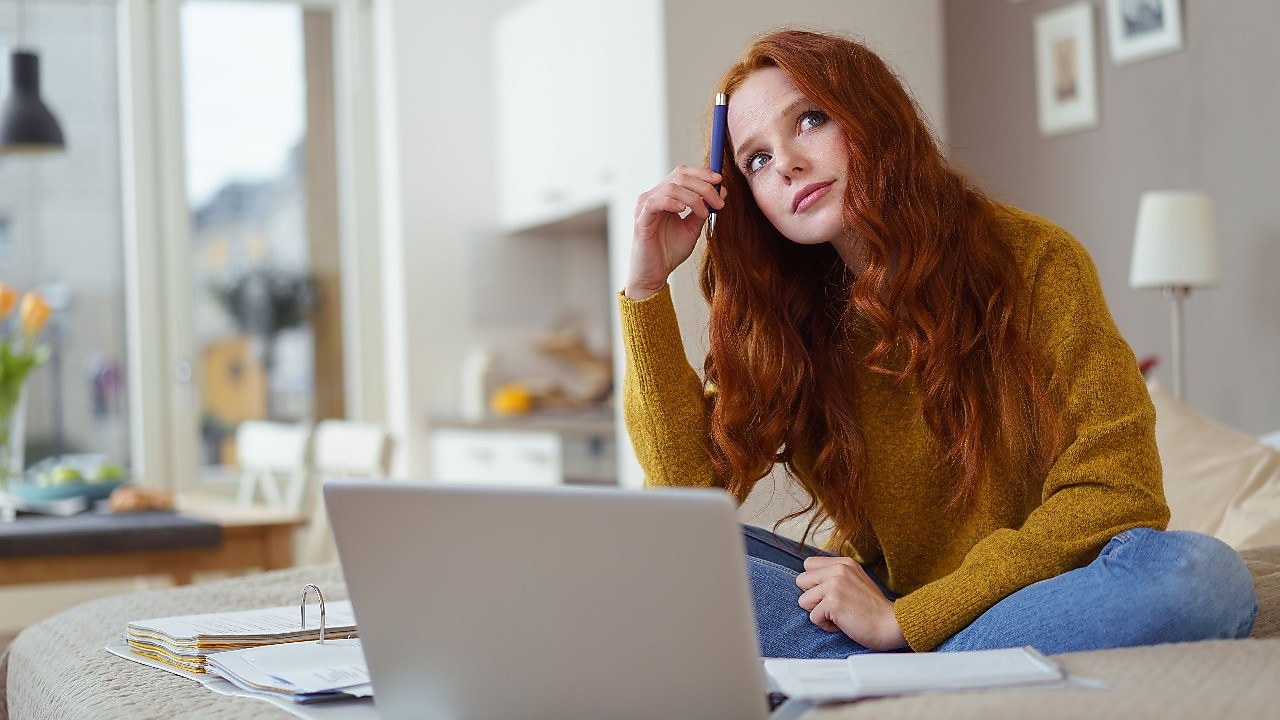 Eine junge Frau sietz in einem hellen Raum vor ihrem Laptop und überlegt.