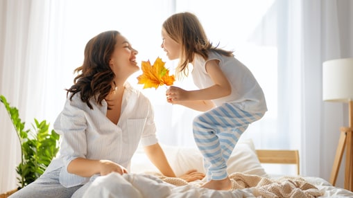 Eine Frau und ein kleines Kind lächeln sich auf einem hellen Bett an; das Kind hält bunte Herbstblätter in der Hand.
