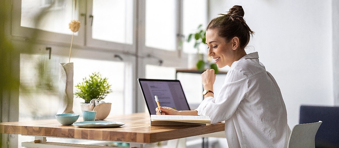 Frau sitzt vor einem Laptop und schreibt auf einen Zettel.
