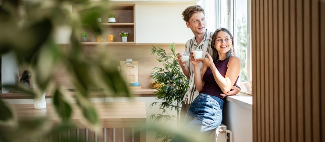 Paar steht mit Getränken am Fenster einer hellen, modernen Küche mit vielen Zimmerpflanzen.