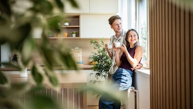 Paar steht mit Getränken am Fenster einer hellen, modernen Küche mit vielen Zimmerpflanzen.
