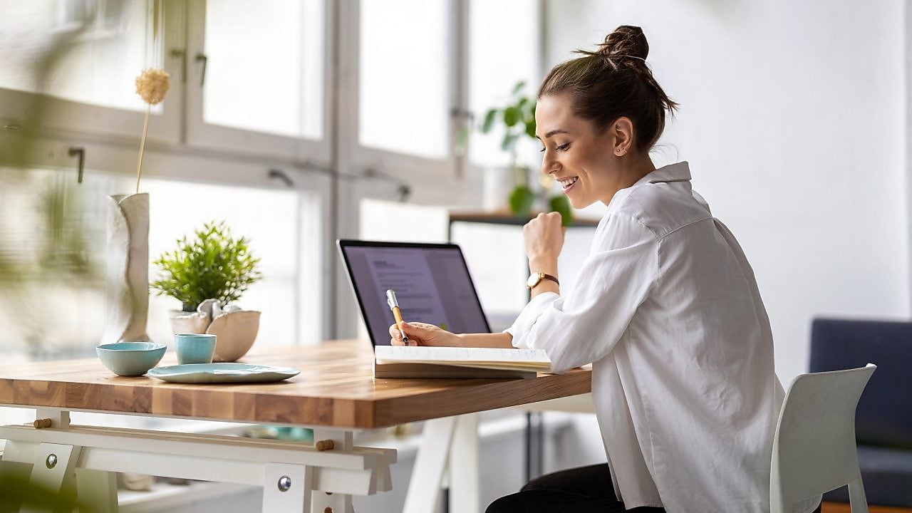 Frau sitzt vor einem Laptop und schreibt auf einen Zettel.