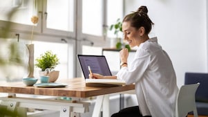Frau sitzt vor einem Laptop und schreibt auf einen Zettel.