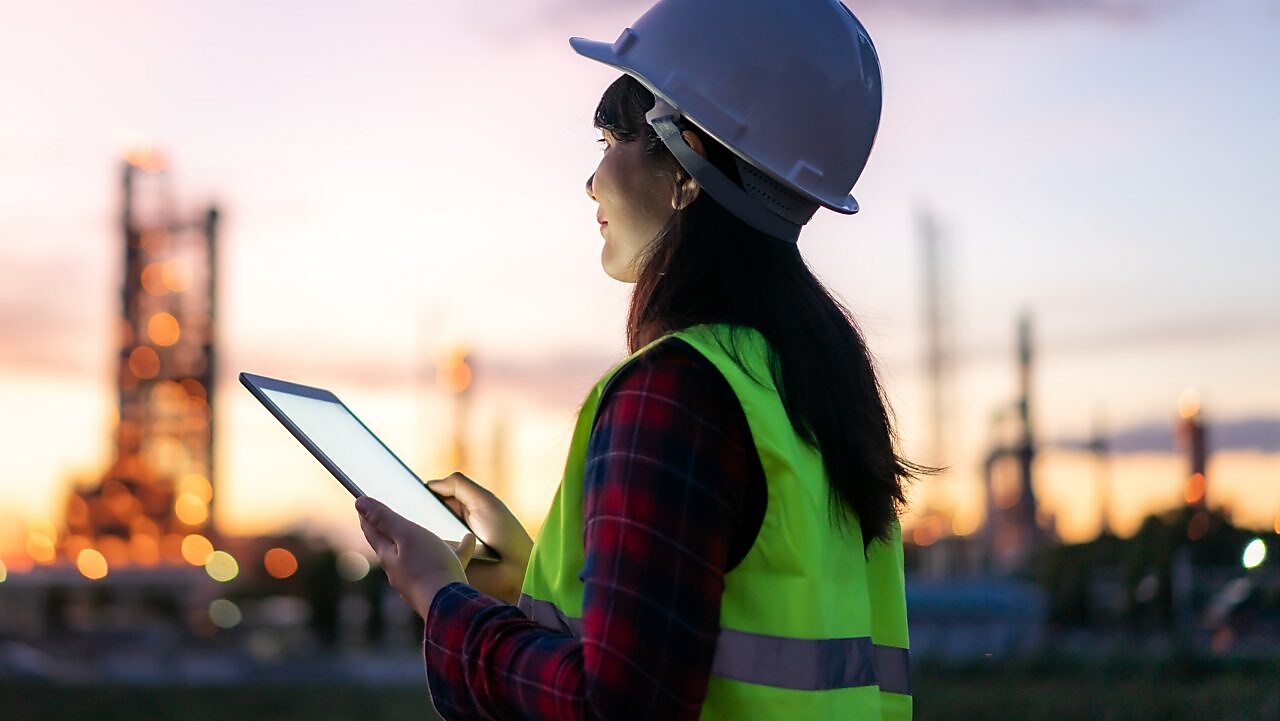 Eine Frau steht bei Sonnenuntergang mit Schutzkleidung und einem Tablet in der Hand im Außenbereich einer Raffinerie.