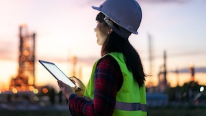 Eine Frau steht bei Sonnenuntergang mit Schutzkleidung und einem Tablet in der Hand im Außenbereich einer Raffinerie.