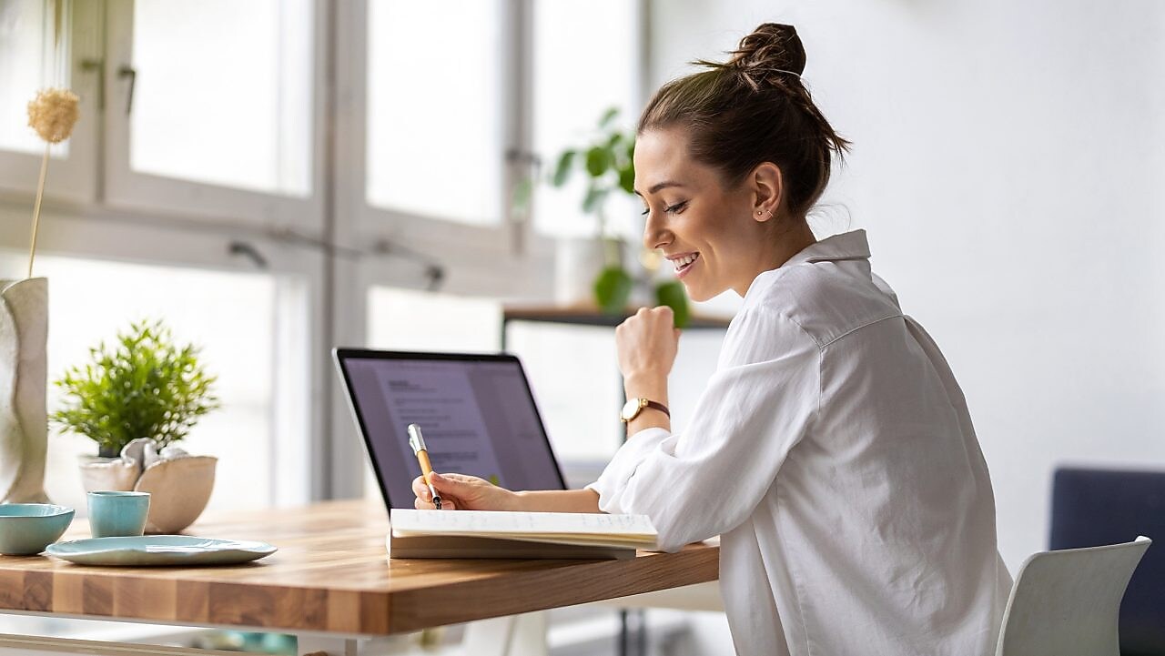 Frau sitzt vor einem Laptop und schreibt auf einen Zettel.