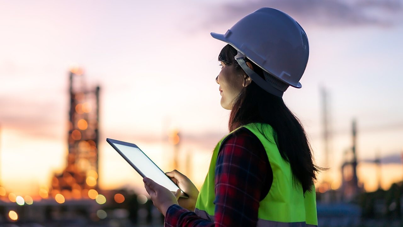 Eine Frau steht bei Sonnenuntergang mit Schutzkleidung und einem Tablet in der Hand im Außenbereich einer Raffinerie.