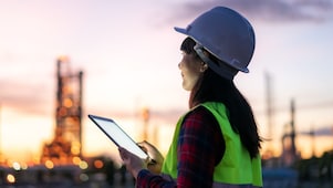 Eine Frau steht bei Sonnenuntergang mit Schutzkleidung und einem Tablet in der Hand im Außenbereich einer Raffinerie.