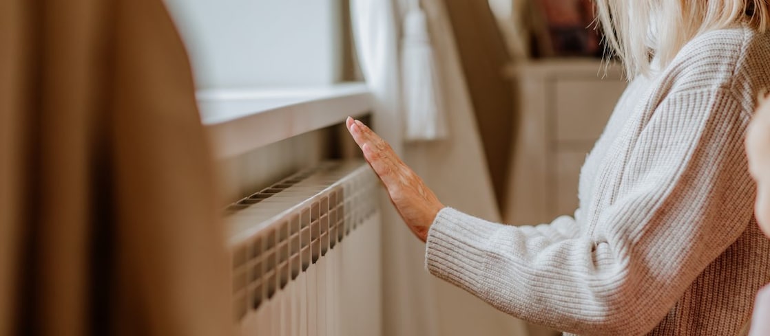 Eine Frau in einem beigen Winterpullover steht in der Nähe der Heizung und wärmt sich die Hände 