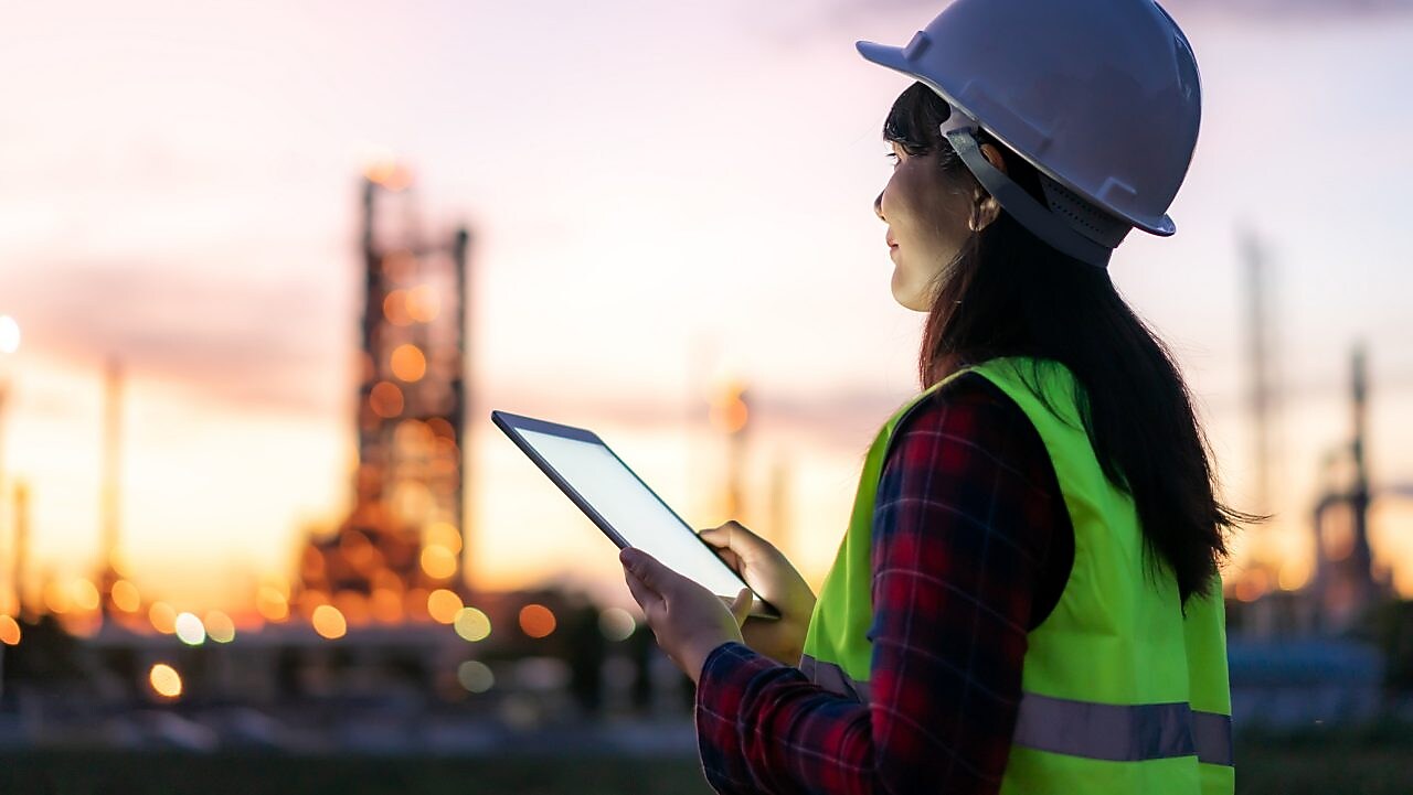 Eine Frau steht bei Sonnenuntergang mit Schutzkleidung und einem Tablet in der Hand im Außenbereich einer Raffinerie.