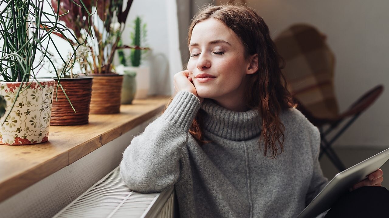 Eine Frau lehnt zufrieden an ihrer Heizung und hält ein Tablet in der Hand.