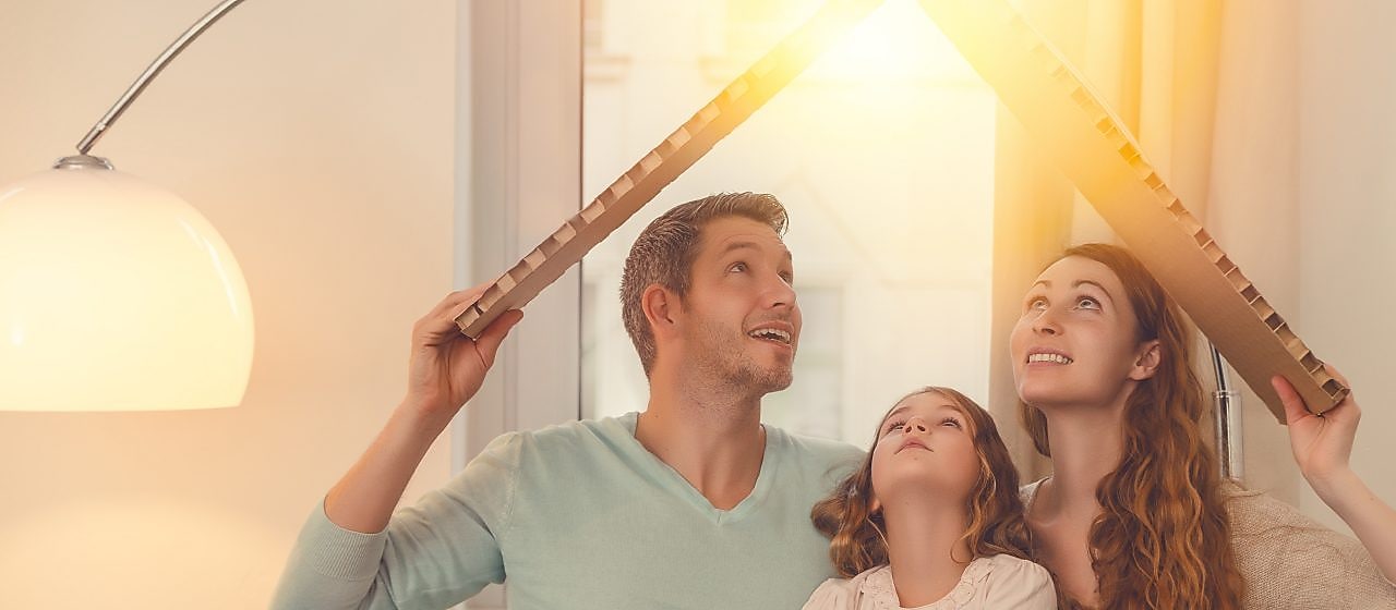 Ein Mann, Frau und ein Kind sitzen in einem Haus, halten sich ein Karton über den Kopf, das aussieht wie ein Dach. Die Sonne scheint hindurch.
