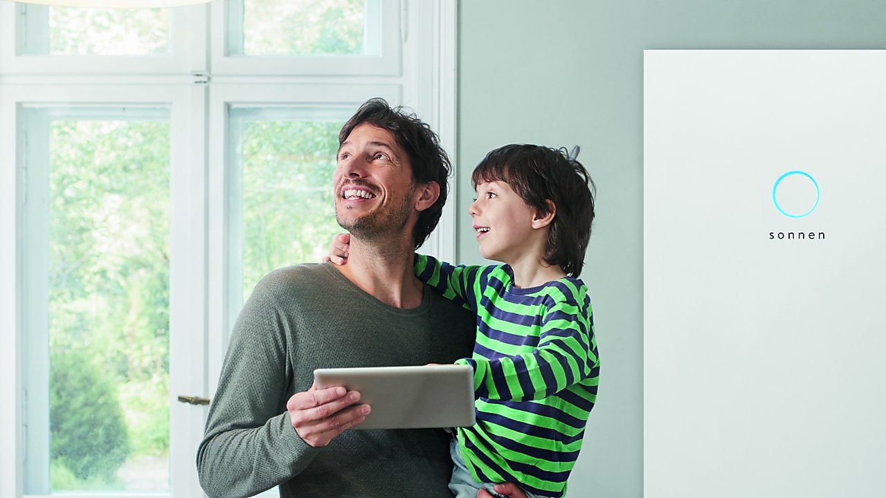 Ein Mann steht mit einem Tablet vor einer sonnenBatterie und hält sein Sohn auf dem Arm.