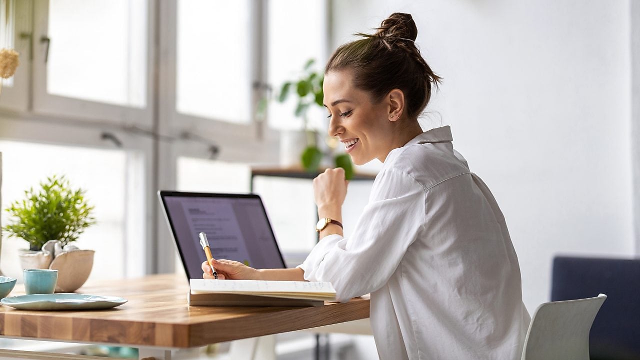 Frau sitzt vor einem Laptop und schreibt auf einen Zettel.