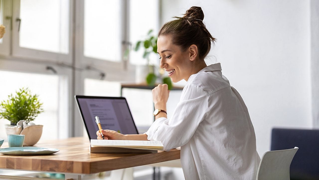 Frau sitzt vor einem Laptop und schreibt auf einen Zettel.