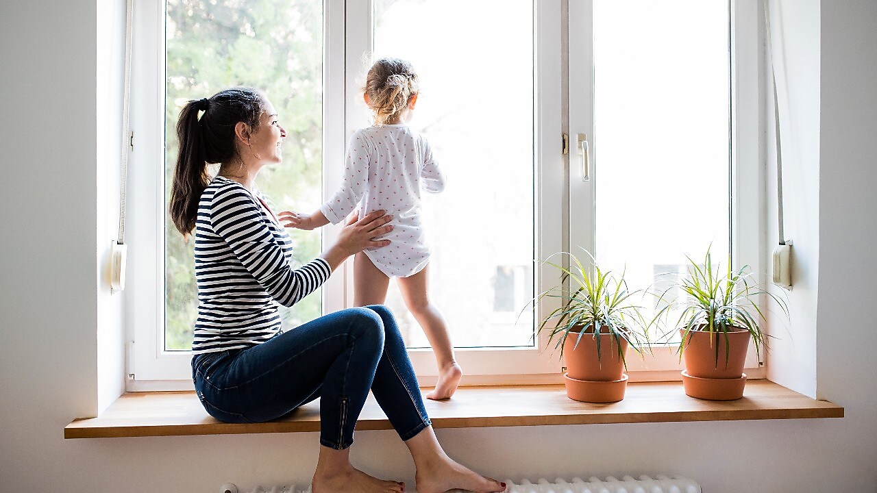 Frau sitzt auf einer Fensterbank und hält ihr Kind mit einer Hand fest, welches auf der Fensterbank sitzt. Beide sind Barfuß.