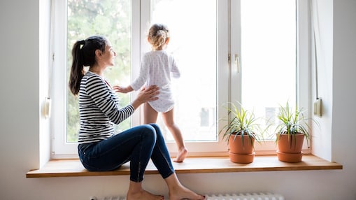 Frau sitzt auf einer Fensterbank und hält ihr Kind mit einer Hand fest, welches auf der Fensterbank sitzt. Beide sind Barfuß.